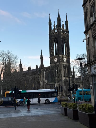 A tall, Gothic church with clock faces dominates the view. Trees stand bare in the foreground, partially obscuring lower portions of the church. A blue and white bus, along with other street vehicles, occupies the street level. Several pedestrians are visible near the bus. The sky is clear and light blue. Buildings border the scene at the top.