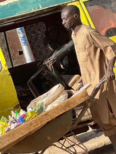 A bald male, dressed in a light brown tunic, pushes a metal wheelbarrow laden with numerous plastic bottles containing various colored liquids and white plastic bags. His right hand grasps the wheelbarrow handle as he moves leftward. In the background, a yellow and green vehicle, likely a bus or large van, displays an open interior with a dark steering wheel, a damaged seat, and a reflective pole inscribed with "HON-SA" in blue text. An obscured individual is visible within the vehicle's interior. The ground is dusty and uneven. The scene is located in Zaria, Nigeria.