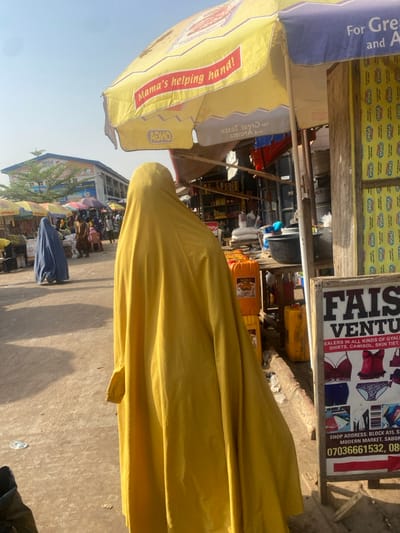 This image depicts a bustling outdoor market scene in Zaria, Nigeria, captured during bright daylight with clear skies, indicated by strong shadows and overhead light. The photograph is oriented vertically, rotated 90 degrees clockwise.
In the foreground, a person is largely obscured, draped in a vibrant yellow garment that covers most of their body. Their head is visible, also covered in yellow fabric. To the left, a large yellow market umbrella is prominent, displaying text such as "Mama's helping hand!" and branding for "ONGA." A blue umbrella is partially visible next to it. These umbrellas provide shade over market stalls.
The background reveals more of the market environment. To the right, a white signboard for "FAISAL VENTURES" advertises "DEALERS IN ALL KINDS OF GYALI, SHIRTS, CAMISOL, SKIN TIET." The sign also provides a "SHOP ADDRESS: BLOCK A15, S MODERN MARKET, SABON" and contact numbers "07036661532, 08". Behind this sign, a wall is covered with repeated "ONGA" branding. Various yellow jerry cans or containers are visible on the ground near the stalls. Further into the distance, other market stalls, goods, and several other people (partially obscured) can be seen, contributing to the lively atmosphere. A distant building with a "MEGA" sign is also visible under the clear sky. The ground is a paved surface, showing some minor debris.