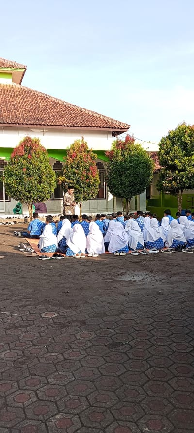 A group of people are sitting on the ground in front of a building in Cangkuang, Indonesia. The people are wearing white headscarves and blue shirts. They are sitting in rows, facing a man standing in front of them. The man is wearing a white headscarf and a blue shirt. There are trees in the background. The ground is paved with hexagonal tiles.
