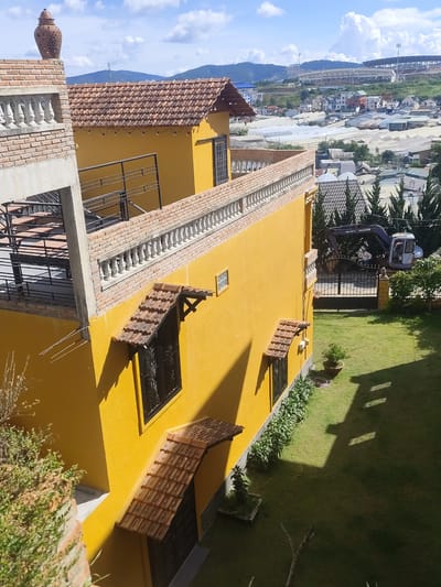 The image shows a yellow building in Lâm Viên - Đà Lạt, Vietnam. The building has a red tiled roof and a brick balcony with a white balustrade. There is a large excavator in the background, parked behind a black gate. A person is visible in the excavator's cabin. The building is surrounded by a green lawn and some trees. In the distance, there are other buildings, greenhouses, and mountains under a partly cloudy sky.