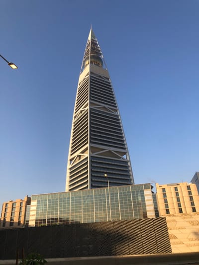 A low-angle shot displays the Kingdom Centre in Riyadh, Saudi Arabia, against a clear blue sky. The building's triangular apex points to the left. Its unique architecture incorporates screened panels. Adjacent to it is a modern building with glass and steel accents, alongside a beige building. A street light pole is visible in the bottom left corner, adding perspective to the building's scale.
