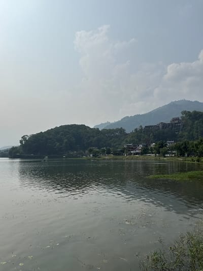 The image shows a view of Phewa Lake in Pokhara, Nepal. The lake is calm with a muted gray color reflecting the cloudy sky. The opposite shore is lined with a lush, green hillside dotted with buildings and trees. The overall scene is tranquil with the blend of natural landscape and human development.
