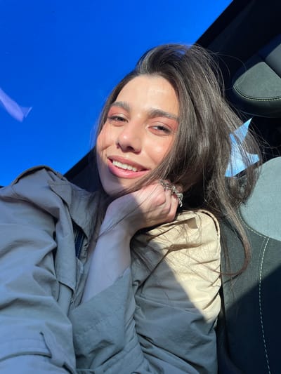 A young woman with long, dark brown hair is captured in a close-up, smiling warmly at the viewer. She appears to be inside a car, possibly lying down or leaning back, with a dark car seat featuring light stitching visible behind her. She wears a light beige or khaki-colored jacket or trench coat. Her left hand, adorned with a silver-toned ring on her ring finger, rests gently near her chin. Direct sunlight illuminates her face, casting noticeable shadows and highlighting her features, including prominent eyebrows and light-colored eyeshadow. The left side of the frame is dominated by a vibrant, clear blue expanse, likely a car window or sunroof reflecting a bright, cloudless sky. The strong sunlight indicates it is daytime with sunny weather. Her expression conveys happiness and relaxation. There is no visible text in the image. The setting is an indoor vehicle environment, with context suggesting the location is Apaga, Armenia.