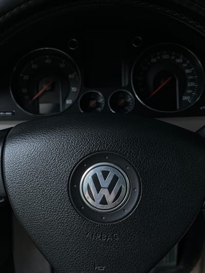 This is a close-up, low-angle shot of a car's interior, focusing on the steering wheel and dashboard. The steering wheel is black and textured, with the Volkswagen logo prominently displayed in the center. The word "AIRBAG" is visible in raised lettering on the steering wheel. To the left of the steering wheel, the dashboard features several gauges with red needles. One large gauge shows speed markings from 0 to 260, with numbers like 80, 100, 140, 180, 200, 220, and 240 visible. Smaller gauges are also present. The overall impression is a view from the driver's seat, suggesting the car is either parked or stationary, possibly in Lviv, Ukraine, given the location context. The lighting is dim, and there are no people visible, emphasizing the object-focused nature of the image. The time of day and weather are not discernible from the image.