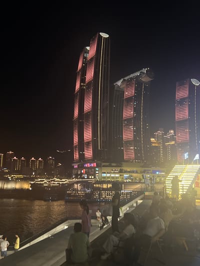 The image captures a nighttime scene along the Yangtze River in Jiangbeicheng Street, Chongqing, China. Dominating the background are several illuminated skyscrapers, prominently featuring the Raffles City Chongqing complex with its distinctive sail-shaped structures and the twin towers with pink lighting. The riverfront is lined with buildings, their lights reflecting on the water. In the foreground, a paved walkway or tiered seating area overlooks the river. Several people are present, some standing and some seated, appearing to be observing the cityscape or a performance. The atmosphere is illuminated by the city lights and the architectural lighting of the buildings.