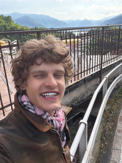 A person with curly blond hair and silver teeth grills is smiling with closed eyes, likely taking a selfie on a balcony. The person is wearing a brown leather jacket over a pink and black checkered scarf. Behind the person are railings and a view of mountains. The location appears to be a balcony in Gittana, Italy. There are metal railings in the foreground and mountains visible in the background. There are tables and chairs visible through the brown railing. A second silver railing is above the brown one.
