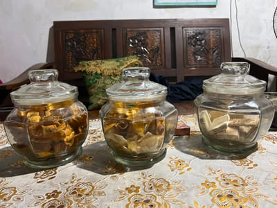 In the city of Layansari, Indonesia, a low-angle, eye-level medium shot captures three glass jars filled with dried food items placed on a patterned tablecloth. The jar on the left contains what appear to be dried fruit slices, predominantly orange-brown in color. The middle jar holds lighter, straw-colored dried food, possibly also fruit or vegetable chips. The jar on the right is filled with rectangular, layered wafers, light brown and white in color.
The jars are positioned in front of a dark wooden bench with carved detailing, suggesting an indoor setting, likely a home. Behind the jars, to the left, a green and gold patterned cushion with fringed edges is visible, resting on the bench. A dark fabric is draped over the bench behind the middle jar. The tablecloth features a repeating pattern of golden flowers and leaves on a light-colored background. The lighting appears to be natural or from an overhead source, casting shadows from the jars onto the tablecloth. The background consists of a plain wall, slightly textured, with a portion of what might be a framed picture or calendar visible at the top. The overall scene is static, with no individuals or discernible actions taking place. The forensic description focuses solely on the visual elements present in the image.