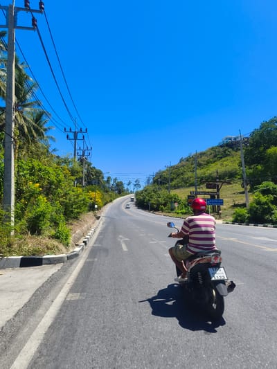 In the city of Baan Thong Po, Thailand, a person wearing a red helmet and a striped shirt is riding a motorcycle on a paved road. The motorcycle's license plate displays Thai characters and the number "1123". The road is lined with lush green vegetation and palm trees, with utility poles and power lines running alongside. In the background, a steep, tree-covered hill rises, with buildings visible at its summit. The sky is a clear, vibrant blue. Several other vehicles are visible on the road in the distance. The overall scene depicts a sunny day on a tropical roadway.