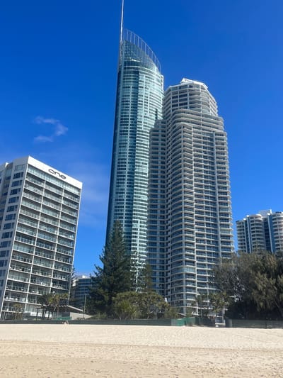 The image depicts a bright, sunny day at a beach in the Gold Coast, Australia. In the foreground, a wide expanse of light-colored sand stretches across the frame. The middle ground features lush green trees and vegetation. Behind the trees, several high-rise buildings dominate the skyline, characteristic of a coastal urban environment.

The buildings are of varying architectural styles and heights. To the left, a predominantly white building with numerous balconies displays the word "one" in large black lettering on its upper facade. Next to it, a very tall, slender skyscraper with a distinctive curved, pointed top and a facade of glass panels rises prominently. To the right of this central skyscraper, a white, multi-tiered building with many balconies is visible. Further in the background, more buildings of similar scale and style can be seen, some with curved balconies and lighter colored facades.

The sky is a clear, vibrant blue with a few wispy white clouds. A single person, appearing as a small figure, is walking on the sand in the lower left portion of the image, near the vegetation line. The overall scene conveys a sense of a modern, developed coastal city with a prominent beach area.