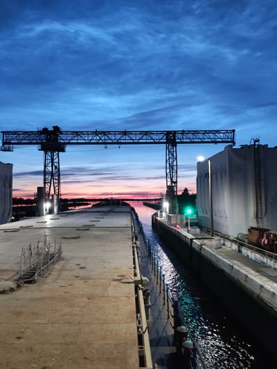 The image depicts a lock on the Sheksna River in Sheksna, Russia, during twilight. A large gantry crane spans the lock entrance, silhouetted against a vibrant sky with hues of deep blue, pink, and orange. The lock chamber itself is filled with dark water reflecting the light from a green traffic signal and overhead lamps. On the right side of the lock, a tall concrete structure, possibly part of the lock mechanism, is visible, with a ladder attached to its side. The left side of the lock features a concrete platform with metal scaffolding and a few visible mooring bollards. The overall scene conveys a sense of industrial infrastructure meeting natural beauty as day transitions to night.