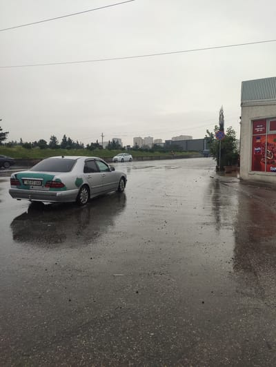 The image depicts an outdoor scene in the city of Motodrom YS, Azerbaijan, characterized by overcast skies and wet ground, suggesting recent rainfall. A silver Mercedes-Benz E-Class sedan, with visible green paint patches on its rear, is parked on the wet asphalt. The car's license plate reads "90-RT-066". In the background, several multi-story buildings are visible, along with trees and streetlights. To the right of the frame, a building with a large window displaying advertisements for Fanta is present. A traffic sign indicating "No Stopping" is also visible. The overall impression is one of a somewhat neglected urban environment under a gloomy sky.