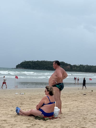 A light blue, overcast sky hangs over a wide, sandy beach in Karon, Thailand. Gentle waves crash on the shore. In the foreground, a woman wearing a blue bikini and blue slippers sits on a patterned mat, her legs crossed. Beside her, a man in patterned swim trunks stands with his hands on his hips. Further down the beach, several people are visible in the shallow water and near the shoreline, including a man in red shorts walking along the wet sand and a man in a dark shirt walking towards the right of the frame. Red flags are planted in the sand along the water's edge. A lush, green island with trees is visible in the background, partially obscured by the ocean.