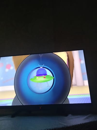 A television screen displays a cartoon image of a green UFO with a purple dome, red and yellow lights, and two antennae. The UFO is suspended within a large blue circle, resembling a magnifying glass or a portal. Behind the blue circle, the blurry background suggests a children's room with pastel-colored walls and toy-like objects. A furry, light brown object, possibly an animal or a stuffed toy, is partially visible in the upper left corner of the screen. The television itself is dark, with a red power indicator light visible on the bottom edge. The screen is positioned on a table covered with a white and grey patterned tablecloth. The overall lighting suggests the room is dimly lit. The image does not contain any identifiable landmarks or features that would indicate its location in Lipetsk, Russia.