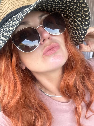 A close-up, waist-up shot shows a woman with reddish-orange hair, wearing a straw hat with a black and white patterned band, and large, dark-lensed sunglasses. She has on a light pink t-shirt and a thin, silver necklace. The woman's lips are painted a light pink color. The background appears to be outdoors, with what looks like a building or structure reflected in her sunglasses. There is no discernible location within the city of Saint-Claude, France.