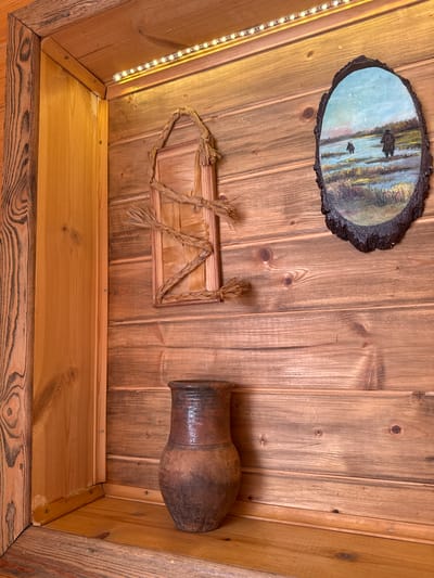 A low-angle shot shows the interior of a wood-paneled structure. In the upper left, there's a framed painting on a slab of wood. Below it, LED strip lights run vertically. To the right of the painting is a framed straw decoration. In the upper right, there is a dark clay pot attached to the paneling. At the bottom of the image, a shelf constructed of light-colored wood is visible. The background consists of brown-toned, aged wood paneling with visible knots. There is no mention of the location in the city of Ketoshi, Russia.