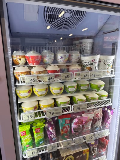 This is a close-up, eye-level shot of a refrigerated display case filled with various ice cream products in a store. The scene is brightly lit by the interior lights of the freezer, and reflections are visible on the glass door. The dominant subjects are the ice cream products themselves, arranged on white wire shelves.

The display includes several brands and types of ice cream. On the left side, there are cylindrical tubs of "Snowy Coconut" and "Coco Cat" ice cream, some featuring cartoonish character designs. Interspersed are individual ice cream cups with colorful lids, also branded "Coco Cat" and featuring similar cute designs, including one with a strawberry motif.

To the right, the shelves are packed with various ice cream bars and possibly more tubs. Prominent among these are green-wrapped "Walls Solero" ice cream bars, a popular brand, and individually wrapped bars with colorful packaging, including purple, pink, and red. Some items have price tags attached, with prices such as "45.00", "79.00", "75.00", "20.00", and "15.00" visible in Thai Baht. Text on packaging and price tags is in Thai and English, with brand names like "Walls", "Ize Coco", and "CocoCat" clearly legible.

The setting appears to be a supermarket or convenience store freezer section, likely in Thailand given the Thai text and currency. The overall impression is of a well-stocked, commercial ice cream display. No people are visible, and the activity is simply the display of frozen goods. The time of day and weather are not discernible.