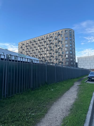 The image shows a modern, curved, multi-story residential building with numerous balconies and windows. The building's facade is constructed with gray brick. To the left of the main building, there is a lower, lighter-colored building with a sign that reads "ШКОЛА ВЕКТОР" (School Vector) in green lettering. A dark gray metal fence runs along the foreground, separating a grassy area with a dirt path from the buildings. A black SUV with a license plate starting with "B811K" is parked on the right side of the frame, adjacent to a sidewalk. The sky above is blue with scattered white clouds. This scene is located in the city of Mytishchi, Russia.