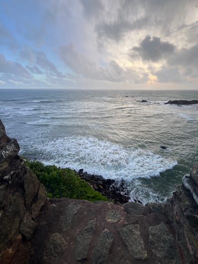 An overcast day at the coast of Galle, Sri Lanka, with dramatic clouds hanging over a choppy sea. The foreground is dominated by rugged, dark rocks, some of which are interspersed with roughly cut stone paving. To the left, a large, dark rock face has graffiti scratched into it, reading "P+S". Green vegetation, a dense cluster of bushes, grows at the base of the cliffs, meeting the dark, rocky shoreline where waves crash. Beyond the immediate coastline, the ocean stretches out, a turbulent expanse of grey-green water with white-capped waves and a few dark rocks emerging from the surface further offshore. The horizon line is blurred by the diffused light and low-hanging clouds. The overall scene is one of raw, natural beauty under a moody sky.