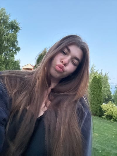 A young woman with long, brown hair is taking a selfie outdoors. She is puckering her lips and winking. She is wearing a dark-colored top. The background shows a blue sky with some clouds, green trees, and a lawn. There is a small wooden structure in the distance. The location is not specified as Мещёры, Russia in the image.