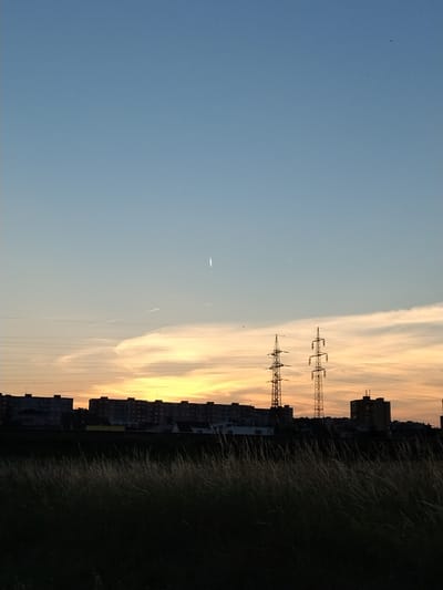 This photograph, taken from the outskirts of Pilsen, Czechia, captures a twilight sky over a residential and industrial landscape. The upper portion of the image is dominated by a clear blue sky that transitions to hues of orange and yellow near the horizon, indicating the setting sun. A faint, white streak, possibly a contrail from an aircraft or a meteor, is visible in the upper sky.
Below the sky, a line of apartment buildings stretches across the middle ground, silhouetted against the sunset. Two tall, lattice-work electricity pylons stand prominently in front of the buildings, with power lines extending between them and horizontally across the scene. A few small birds are discernible in the sky.
The foreground is filled with tall, dry grass and vegetation, rendered in dark silhouette against the brighter background. The overall impression is one of an urban fringe at dusk.