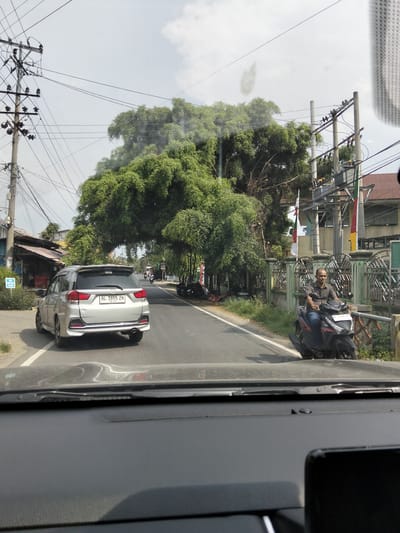 The image captures a street scene in Keudematangglumpangdua, Indonesia, on a cloudy day. In the foreground, the dashboard of a vehicle is visible, suggesting the photo was taken from inside a car. Ahead, a silver multi-purpose vehicle (MPV) with license plate "BL 1695 ZH" drives on the asphalt road. To the right of the MPV, a man wearing a gray shirt and blue jeans rides a black scooter. He is looking towards the left side of the frame and appears to be smiling. Behind the man, a green metal fence runs along the side of the road, bordering a property with a building featuring a red tiled roof. Large, dense trees with sprawling branches overhang the road, creating a canopy effect. Utility poles with numerous wires are prominent on both sides of the street. Further down the road, other vehicles and people are visible, indicating moderate street activity. A small market or shop with a weathered roof is situated on the left side of the road, near the silver MPV.