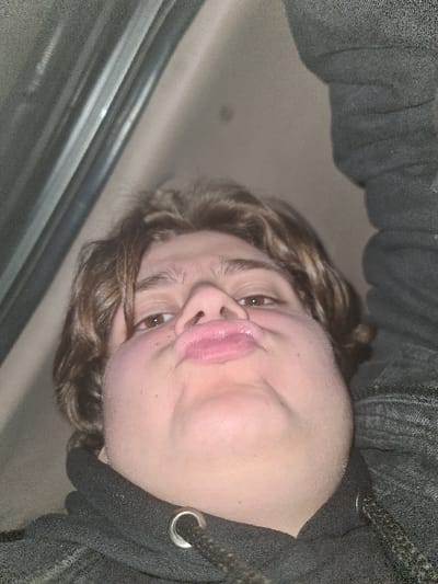 A close-up selfie features a person with light skin, brown eyes, and light brown curly hair. They are wearing a dark gray hooded sweatshirt with the drawstring visible. The camera angle is tilted, causing a distorted perspective, particularly noticeable in the stretched facial features and pursed lips. A gray surface is partially visible on the left side of the frame. The overall lighting is dim.
