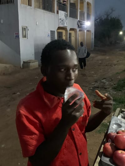 This portrait-oriented photograph, rotated 90 degrees clockwise, depicts an outdoor scene at dusk or night, likely in an urban or semi-urban area consistent with Ilorin, Nigeria.

The central subject is a young person with dark skin and short, dark hair, wearing a prominent red puffer jacket. They are looking slightly upwards and to their left (relative to their body), with a clear plastic object held near their mouth, suggesting they are drinking or eating. Their right hand is raised, with the index finger extended in a pointing or gestural motion.

To the right of the frame, a small market stall or table is visible, displaying a tray piled with red apples, some of which are contained within clear plastic bags. The ground beneath the figures and the stall is unpaved and appears to be dirt or earth.

In the background, on the left side of the frame, multi-story buildings with visible windows and balconies are present. Artificial lights from within or around these structures illuminate parts of the scene. Another person, wearing a light-colored hooded top and dark trousers, can be seen in the distance, near the buildings. A faint sign with the letters "POS" is visible on one of the background buildings. The overall impression is a casual evening street scene, possibly centered around a vendor.