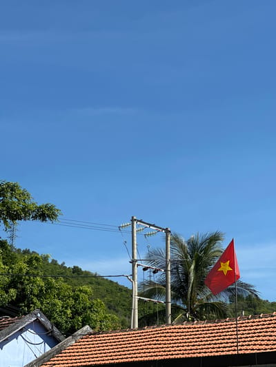 The image captures a bright, clear blue sky over a rooftop in Ninh Ích, Vietnam. A red flag with a yellow star, the flag of Vietnam, is prominently displayed on a pole extending from the rooftop. The rooftop is covered with orange terracotta tiles. Behind the flag, a palm tree with lush fronds is visible. To the left, power lines stretch across the frame, supported by utility poles, and a dense green hillside rises in the background. A portion of a light blue building is visible on the left side of the image, with a portion of a metal tower also visible in the far distance on the left. The scene is tranquil and suggests a sunny day in a coastal or hilly region of Vietnam.