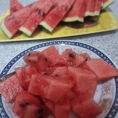 This media file shows a close-up, overhead view of a plate of sliced watermelon and a yellow tray also filled with watermelon slices. The watermelon is cut into bite-sized cubes and irregular triangular pieces. Some pieces still have black seeds visible. The watermelon is a vibrant red color with a thin white rind and a green outer peel on some slices.
The watermelon slices are arranged on two surfaces. The left side of the image features a yellow tray with several triangular slices of watermelon. The right side of the image displays a white plate with a decorative blue and white patterned rim, filled with cubes and larger chunks of watermelon. A silver fork is partially visible on the plate, suggesting the watermelon is ready to be eaten.
The background appears to be a speckled countertop, possibly in a kitchen or dining area. The lighting suggests it is daytime, likely indoors. There is no visible text in the image. The scene depicts a simple, refreshing snack, likely prepared for enjoyment. The context of "Dunnae-myeon, South Korea" suggests a domestic setting in this region.