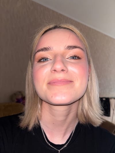 The media file is a close-up, rotated portrait of a young Caucasian woman with fair skin and blonde hair. She is wearing a black top and a silver necklace. Her makeup includes blush on her cheeks and mascara on her eyelashes. She is looking towards the camera with a slight smile. The background is blurred and appears to be an indoor setting with neutral-colored walls and floor. The lighting suggests it is daytime or well-lit indoors. There is no visible text.