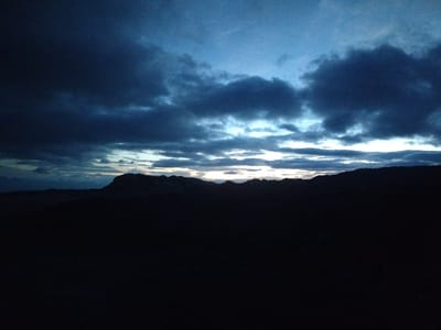 The image depicts a twilight scene of a mountain range with a dark, overcast sky. The silhouette of rugged mountains is visible against a backdrop of deep blue and grey clouds, with streaks of pale light breaking through near the horizon. The foreground is entirely consumed by shadow, rendering any details indistinguishable. The scene appears to be set in a natural, undeveloped landscape, likely a remote area, given the lack of any man-made structures or signs of human activity. No people, subjects, or objects are discernible. The location in the city of Naminga, Russia is not apparent from the visual information provided.