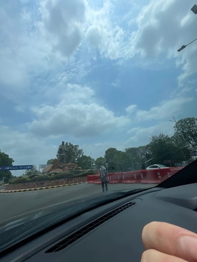 The image depicts a view from inside a vehicle, looking out at an outdoor scene. In the distance, a large bronze statue of a standing man is visible, positioned atop a rocky and landscaped base. The statue is set against a backdrop of lush green trees and a partly cloudy sky.
In the mid-ground, there is a road with traffic barriers. A person wearing a light-colored shirt and dark pants, and possibly a mask, is standing near the barriers. A white car is also visible on the road. Road signs indicating "T1 Departures" and "T1 Arrivals" are present on the left side of the image, suggesting the location is near an airport. The foreground shows the dashboard of the vehicle from which the photo is taken, with a person's thumb visible on the right.
The scene is set in Ahmedabad, India, with the statue likely being a prominent landmark within the city. The overall impression is a daytime urban environment with clear, cloudy skies.