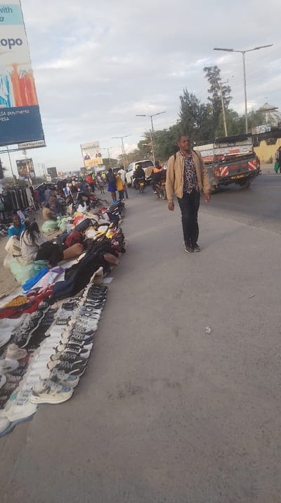 The image captures a bustling street market scene alongside a busy road, likely in Oloosirkon/Sholinke ward, Kenya. In the foreground, a man wearing a light brown jacket and dark pants walks along the side of the road, looking towards the left. To his left, a long line of informal market stalls stretches along the pavement, displaying various goods.
The stalls are primarily comprised of clothes and numerous pairs of shoes laid out on cloths or mats directly on the ground. Several people, including vendors and potential shoppers, are present at these stalls; some are seated on the ground attending to their merchandise, while others appear to be browsing. One person is wearing a light blue top, and a white bag nearby has "THINKING LUCK?" written on it.
On the right side of the image, a paved road accommodates moving vehicles, including a white pickup truck with a utility bed, partially obscured cars, and motorbikes. The pickup truck has "TOY TA" visible on its tailgate, likely part of "TOYOTA," and a Kenyan license plate is partially visible.
The scene suggests late afternoon or early evening, characterized by soft, diffused light and an overcast sky with scattered clouds. Streetlights are visible along the road, indicating the approach of dusk or a generally cloudy day. In the background, large billboards advertise services; one prominent sign on the far left reads "kopo" and "ESA payments PESAT," strongly suggesting an M-PESA mobile money advertisement. Another billboard further down the road displays "CROSSOVER." The overall impression is that of a lively commercial area.