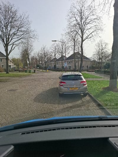 The image presents a static daytime scene in a residential area of Teteringen, Netherlands, viewed from within a vehicle, with its blue dashboard and a portion of the windshield occupying the lower foreground. The sky above is clear, and tree shadows are cast upon the pavement, indicating sunlight.
In the midground, a silver or champagne-colored hatchback vehicle, bearing Dutch license plate "57-TJK-1," is parked with its rear angled towards the right. It rests on a paved surface composed of light-colored interlocking pavers. To the immediate right of the parked vehicle is a narrow strip of green grass, bordered by a curb. A large, bare-branched tree trunk is partially visible on the far right edge of the image, obscuring part of the background.
The background shows a residential street extending into the distance. It is lined on both sides by two-story houses featuring light-colored facades and dark-pitched roofs. Numerous bare-branched deciduous trees are prominent throughout the scene, flanking the street and interspersed between residences. A streetlamp with a horizontal arm is positioned over the road on the right side. On the left side of the street, two green trash bins are visible near some trees, along with a partial view of a dark-colored vehicle further down the road. A blue and white sign, possibly indicating a bus stop, is visible further down the street on the right. No individuals, overt actions, or interactions are depicted within the frame.