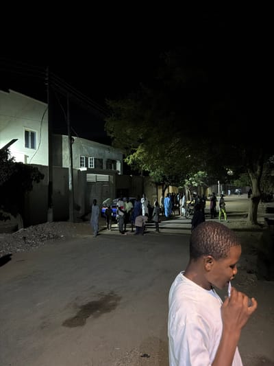 The scene depicts a nighttime street in Gandu Albasa, Nigeria. In the immediate foreground, to the right, a young male with short, dark hair, wearing a white t-shirt, is visible from the chest up, facing left. He is holding a white object, consistent with a toothbrush, to his mouth, suggesting oral hygiene. The ground directly in front of him is an unpaved dirt surface with a dark, irregular liquid stain.
In the midground, an unpaved road extends into the distance, illuminated by artificial light sources. On the left side of the road, a pile of dark debris or rubble is present. Beyond this, a two-story building with a light-colored, possibly white or cream, facade and a visible window is illuminated. Adjacent to it, a utility pole with overhead wires stands. To the right of the pole, another building with a grey facade is visible, featuring a horizontal-slatted metal gate and two windows with white frames on its upper story.
A group of approximately 15-20 individuals, predominantly adult males, are gathered on the road between the buildings and trees. Many are dressed in traditional robes of various colors, including blue, grey, and white. A blue vehicle is partially obscured by the group. Most individuals are standing, appearing to engage in casual conversation or observation. One person on the far left of the group wears a light grey robe. Further down the street, to the right, a person is seen with a bicycle.
Trees with visible foliage line the right side of the street and extend into the background, with some leaves illuminated by ambient light. The sky is dark, indicating night. Faint lights from distant buildings or streetlights are visible further down the road, contributing to the overall illumination of the scene.