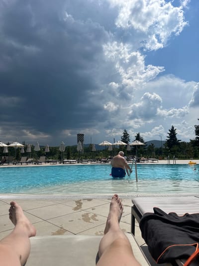 The image depicts a poolside scene with a person entering the water. The perspective is from someone sitting on the edge of the pool, with their legs and feet visible in the foreground. The person in the water is a man wearing blue swim trunks and is holding onto the pool ladder as he begins to descend.
The setting is an outdoor swimming pool with clear blue water and a tiled surrounding area. Several lounge chairs and umbrellas are arranged along the pool deck, suggesting a resort or leisure facility. In the background, a line of trees and mountains can be seen under a dramatic sky filled with both bright white, puffy clouds and darker, more ominous storm clouds. This contrast suggests a transition in weather.
The time of day appears to be daytime, given the sunlight illuminating the scene, but the dark storm clouds indicate a potential for rain or a change in weather. The overall mood is one of relaxation and leisure, albeit with a hint of impending weather change. There is no visible text in the image. The location is identified as Kazanlak, Bulgaria.