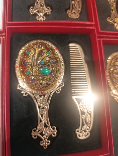 This image displays a collection of ornate, antique-style vanity items, likely a mirror and comb set, presented in a red velvet-lined box. The mirror is circular with a highly decorative, gold-toned frame encrusted with small, colorful gemstones. The handle of the mirror is intricately carved with floral motifs. To the right of the mirror lies a matching comb, also with a decorative handle. The items are arranged in individual compartments within the box, suggesting they are part of a set. The background is dark, likely black velvet, which contrasts with the golden hues of the vanity items and the vibrant red of the box. The lighting suggests an indoor setting, possibly a display case or retail environment. This image does not contain any elements that would allow for the determination of its location in Dub, Montenegro.