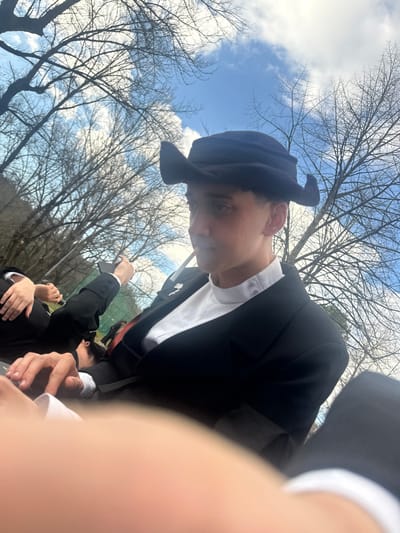 A low-angle, outdoor photograph captures a young individual, likely male, positioned in the foreground, appearing seated or reclining. The subject is wearing a black suit jacket over a white, high-collared shirt, and a distinctive black hat with two prominent upward-curving flaps on its sides. A metallic pin is affixed to the left lapel of the jacket. A thin, cylindrical object, possibly a cigarette or pen, is held near the subject's mouth, pointing upwards. The individual's gaze is directed slightly downward and to their left.
To the left of the main subject, a second individual is partially visible, also dressed in a black suit and white shirt, holding a dark, rectangular object (possibly a smartphone). Further left, components of a third individual are discernible. The bottom right foreground is blurred, showing a light-skinned hand with a white shirt cuff.
The environment consists of a clear blue sky with scattered white cumulus clouds overhead. Numerous bare deciduous trees with intricate branch patterns dominate the background, indicating a non-summer season. A green net, possibly part of a sports facility, is faintly visible in the mid-ground. The attire worn by the individuals strongly suggests a Portuguese academic "traje," situating the scene within student traditions, likely "praxe académica," in Guimarães, Portugal, possibly within a park or outdoor area near the University of Minho campus.