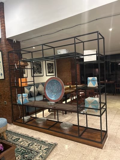 This image depicts the interior of a retail space, possibly a furniture or home decor store, located in Sukawati, Indonesia. The scene is dominated by a large, black metal shelving unit filled with various decorative items. On the shelves, there are patterned bowls, woven baskets, ornate decorative plates, upholstered cubes with geometric patterns, and a stack of orange boxes. Below the shelving unit, a wooden base holds several silver decorative boxes and lids.
In the background, a brick wall is visible, and a blue sofa with striped pillows is situated behind the shelving unit. A dining table with chairs can be seen further in the background, suggesting a potential lounge or cafe area within the establishment. The floor is tiled with a light-colored, geometric pattern. A partially visible rug with intricate green and cream designs lies on the floor in the foreground, next to a wooden table and a blue and tan woven ottoman. The lighting suggests an indoor, possibly commercial setting. There are no people visible in the image.
