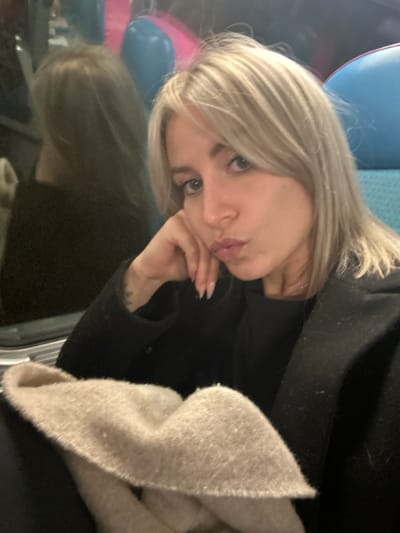 A blonde woman rests her head on her hand. She wears a black jacket. A beige scarf is visible. Her fingernails are manicured. A reflection of her face is visible in the lower-left corner. Blue and pink balloons are in the background, partially obscured. She is looking at the camera.