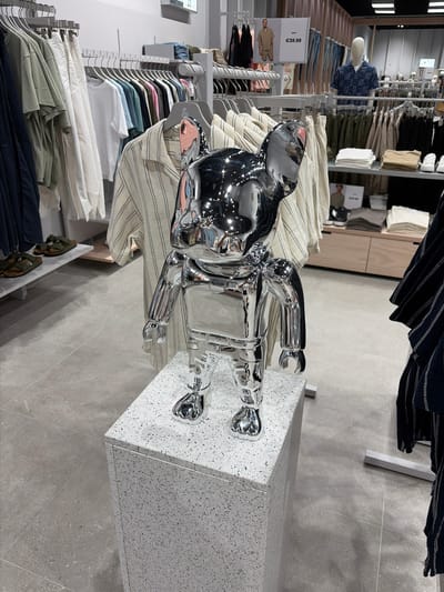 This image shows a retail clothing store interior, likely in Marousi, Greece. The scene is dominated by racks of clothing, primarily shirts and jackets, displayed in a well-organized manner. In the foreground, a striking chrome statue of a human figure stands on a speckled, light-colored pedestal. The statue is highly reflective, mirroring the surrounding environment. Behind the statue, a mannequin dressed in a patterned blue shirt is visible, along with more clothing displays. The overall impression is that of a modern and clean retail space, possibly during daytime operating hours. There are price tags visible, one with "€39.99" clearly legible, indicating a commercial environment. The lighting appears to be artificial, consistent with an indoor retail setting.