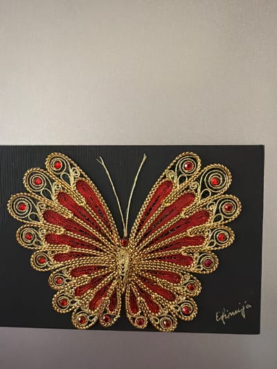 A close-up, eye-level shot features a decorative butterfly artwork mounted on a black rectangular background. The butterfly is intricately crafted from red and gold threads, adorned with small, red, gem-like embellishments. The artwork is signed in script in the bottom right corner. The background is a textured black cardstock. The artwork is set against a plain, neutral-colored wall, with the top portion of the wall being a lighter shade and the bottom a darker shade. The lighting is even, with no distinct shadows. This artwork is located in Pazardzhik, Bulgaria.