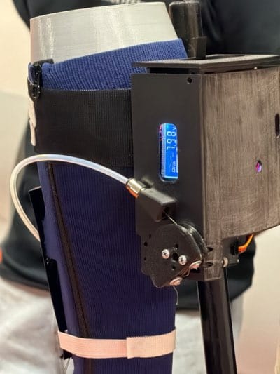 Here is a detailed description of the image:
The image shows a close-up view of a prosthetic leg. The upper portion of the prosthesis is white and appears to be made of a 3D-printed material. Beneath that is a navy blue sock with a black strap securing it. There's a black apparatus attached to the side of the prosthetic leg, which features a small blue display showing digital readings, a clear tube connected to a fitting, and a black pulley mechanism. The apparatus is mounted on a dark wood-like panel. The prosthesis appears to be attached to a metal post. In the background, a person wearing a black shirt with an orange logo is partially visible. The setting appears to be indoors, likely at an exhibition or demonstration area, given the focus on the detailed workings of the prosthetic device. There is no indication of the city of Rochester, United States in the image.