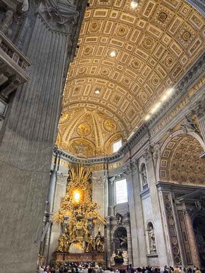 The image displays the interior of St. Peter's Basilica, located in Città del Vaticano, Vatican City, specifically focusing on the apse and the **Cathedra Petri**. The perspective is a low-angle, wide shot looking towards the main altar area.
In the foreground, a large group of people is visible, mostly from the chest up, positioned in rows and facing away from the camera, towards the central monument. They appear to be observing or attending an event. Their clothing is diverse and casual.
The central and dominant feature is the monumental **Cathedra Petri**, an ornate gilded Baroque sculpture. It features a bright circular stained-glass window or illuminated oculus at its center, depicting a dove (symbolizing the Holy Spirit) surrounded by rays of light, clouds, and numerous cherubic figures. Below this, the bronze chair is flanked by four large golden statues of Doctors of the Church. The entire structure is exceptionally gilded, reflecting significant light.
Massive fluted grey stone columns rise vertically on the left side of the frame. On the right, the apse walls are adorned with various architectural elements, including pilasters, niches containing statues, and sections of patterned marble. A large rectangular window with natural light is visible on the upper right, and another lower on the left side of the apse.
The ceiling above is a grand barrel vault leading into a dome, meticulously decorated with intricate gilded coffering. The patterns consist of geometric shapes, including rectangles and octagons, filled with elaborate rosettes and sculptural details in gold and off-white. A frieze with large Greek inscription runs along the top of the apse walls, below the ceiling. Artificial lights are visible along the ceiling, supplementing the natural light. The overall impression is one of immense scale, rich ornamentation, and dramatic lighting.