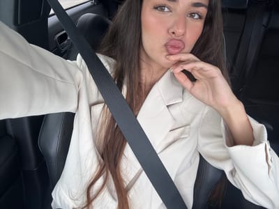 A woman is in a car. She is wearing a white blazer and a black seatbelt. She is making a kissing face with her hand near her mouth. The interior of the car is black leather. The location is unknown and cannot be determined from the image.