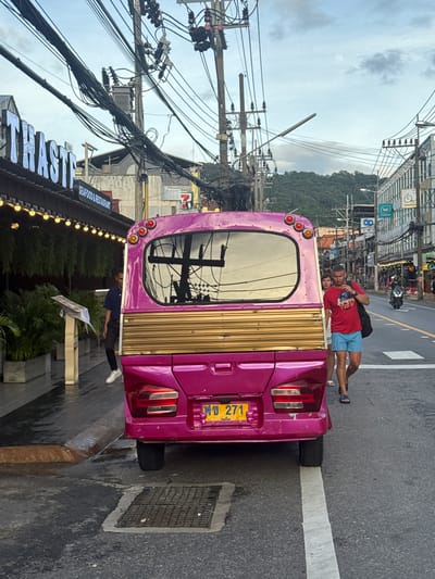 A man and a woman are leaning against the back of a bright pink vehicle, possibly a tuk-tuk, on a street in Karon, Thailand. The man is wearing a red t-shirt and blue shorts, and the woman is wearing a dark blue shirt and black pants. They appear relaxed and are looking at the camera.

The vehicle has a distinctive gold paneling on the rear. A yellow license plate with Thai script and the number "271" is visible. The street is paved and appears wet, suggesting recent rain or a damp environment. Overhead, a complex network of electrical wires and poles dominates the sky, a common sight in many parts of Thailand. In the background, buildings with signs, including one that reads "SEAFOOD & RESTAURANT," and lush green foliage can be seen, indicating an urban or tourist area. The sky is overcast, suggesting it might be daytime, possibly late afternoon or early morning. The overall mood seems casual and relaxed, with the couple enjoying a moment in a bustling street environment.