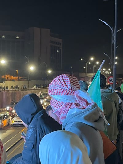 A nighttime gathering of people is visible. Some individuals are draped in traditional red and white patterned cloths. A green flag on a pole is visible. The background includes buildings and streetlights. There are several people wearing hooded jackets.