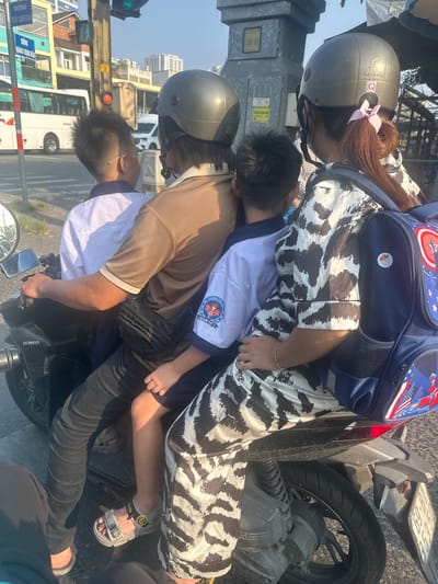 A motorcycle is carrying three individuals on a city street in Ho Chi Minh City, Vietnam. The driver is an adult wearing a brown shirt, black jeans, black sandals, and a gray helmet. Two children are riding behind the driver, with the younger child sandwiched between the driver and an adult wearing a white and brown patterned shirt and a gray helmet. The younger child has on sandals and a school uniform. The adult in the back is wearing a backpack with a Captain America design. The motorcycle, identified as a Honda, is situated near a traffic light. Buildings are visible in the background. The license plate of the motorbike reads 59-L8 181.69. Street signs are also visible, including one that reads "DUONG TRAN XUAN SOAN." Another sign reads "TRUONG TIEU HOC HAM TU," and contains the symbol of the school and its name.