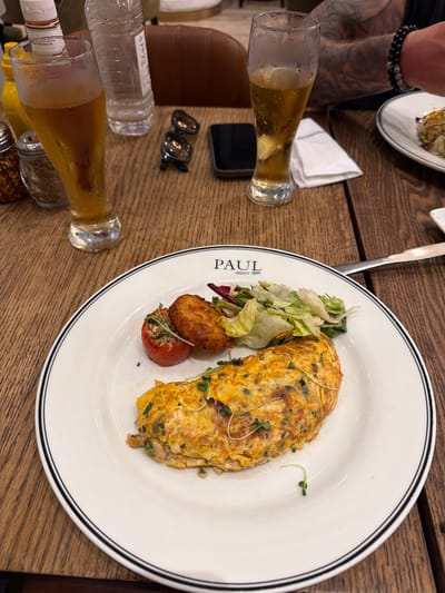 A close-up, overhead shot shows a meal on a wooden table. In the foreground, a glass of amber-colored beverage, likely beer, sits on the table, its surface covered in condensation. Next to it, another glass of the same beverage is partially visible. A clear plastic bottle of water and a bottle of liquid with a white label are also present. Further back, a person's arm, adorned with a black and grey beaded bracelet, is visible, resting on the table. On the table, a white plate with a black rim holds an omelet, a small pile of salad, a fried item, and a stuffed tomato. The word "PAUL" is printed on the plate, along with the text "depuis 1889". In the background, a white napkin is folded. The lighting suggests it is daytime, and the overall scene depicts a casual dining experience.